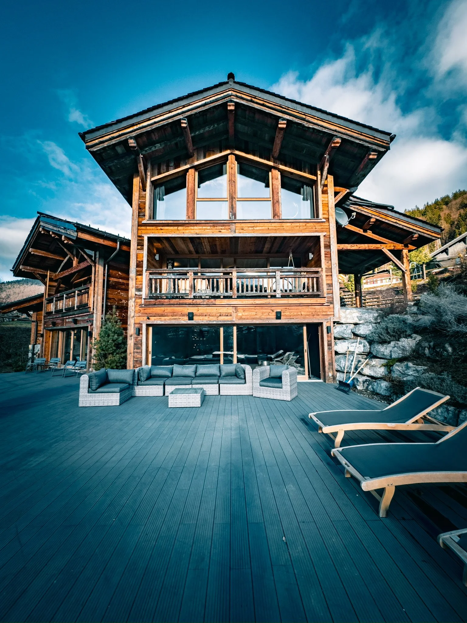 A large wooden house with a spacious deck, wicker outdoor furniture, and lounge chairs. Mountain landscape in the background under a blue sky with some clouds.