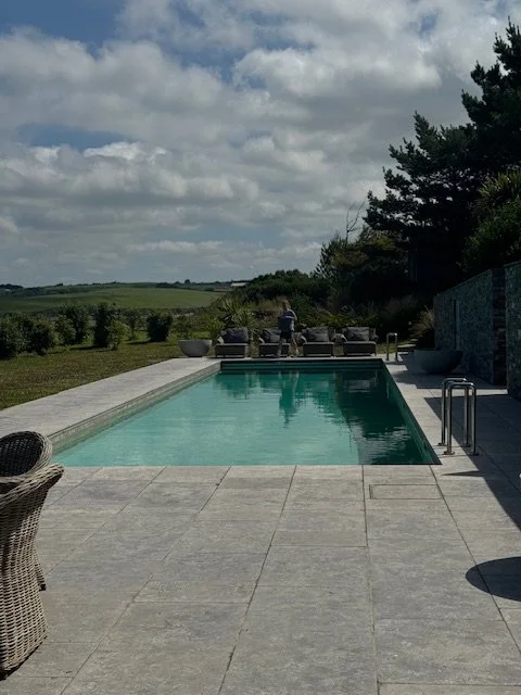 An outdoor swimming pool with a concrete deck, surrounded by trees and grassy fields under a partly cloudy sky.