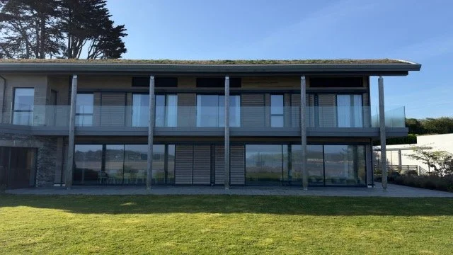 Modern two-story house with large glass windows and a balcony, surrounded by a grassy lawn under a clear blue sky.