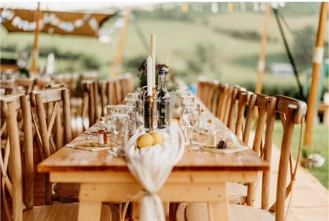 Long outdoor table set for a gathering with wooden chairs, candles, glasses, and a centerpiece with lemons under a tent with string lights visible.