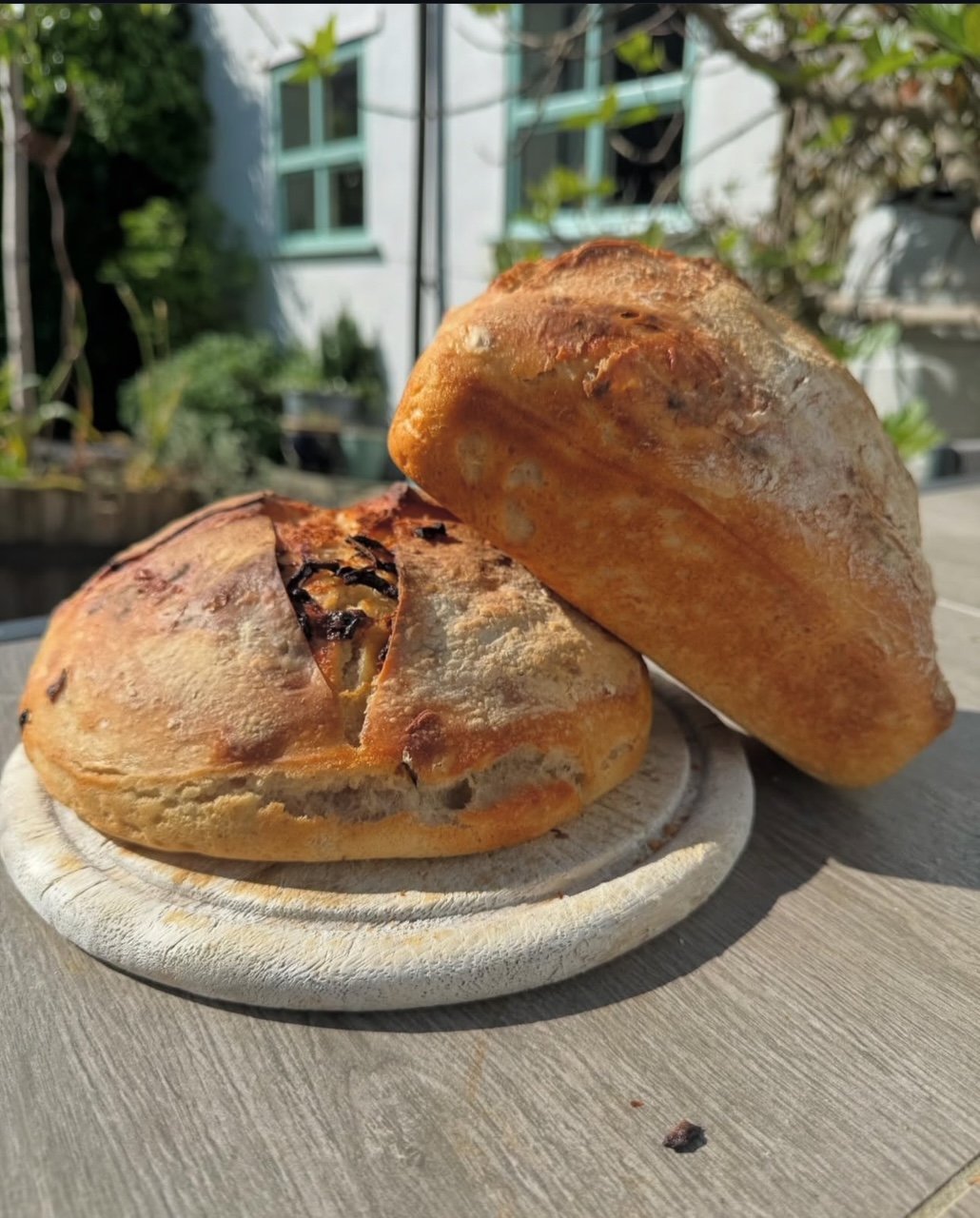 Two rustic bread rolls on a wooden board outdoors, with a garden and a house in the background.
