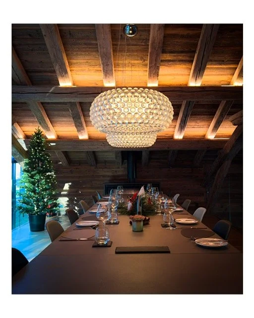 A decorated dining table with place settings in a rustic, wood-beamed room featuring a large, round, chandelier and a decorated Christmas tree.