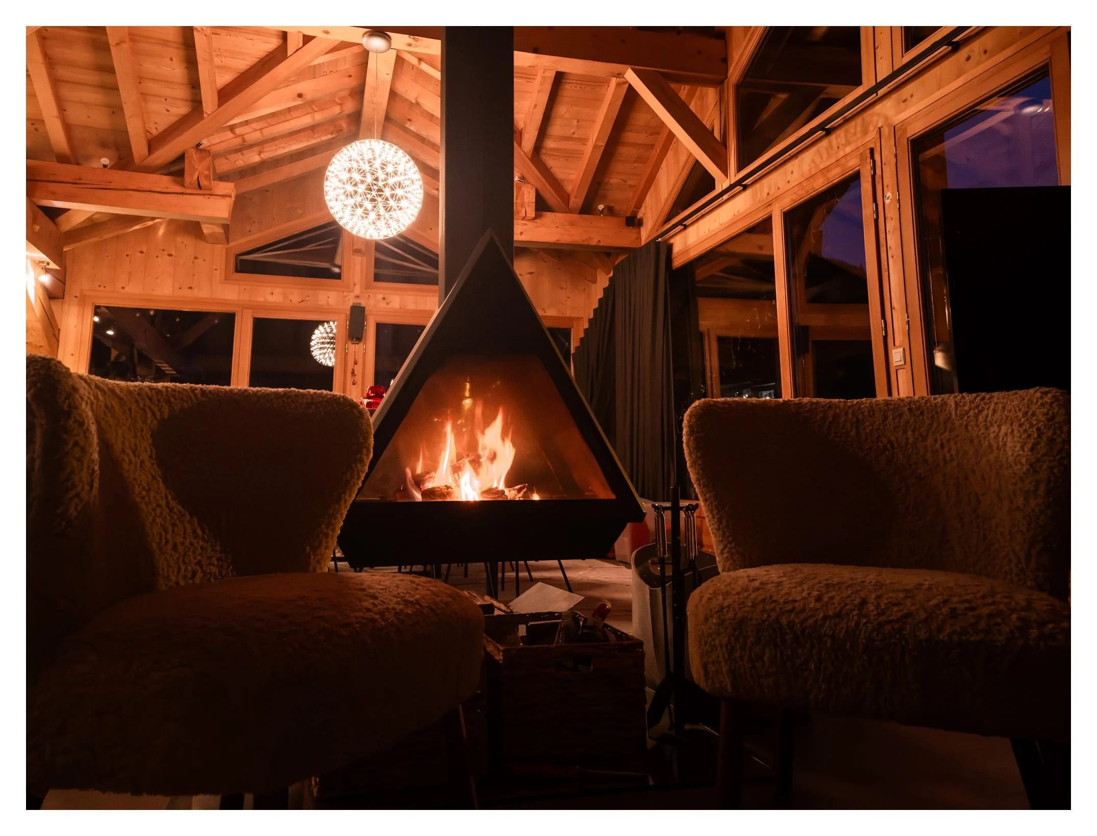 Cozy living room with a central fireplace, two armchairs with textured fabric, large wooden windows, and a wooden ceiling with exposed beams illuminated by a spherical pendant light.