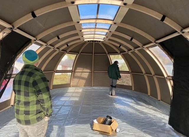 Two people inside a partially constructed dome-shaped structure with a transparent roof, with one person standing near the window and the other looking out, and a cardboard box with tools on the floor.