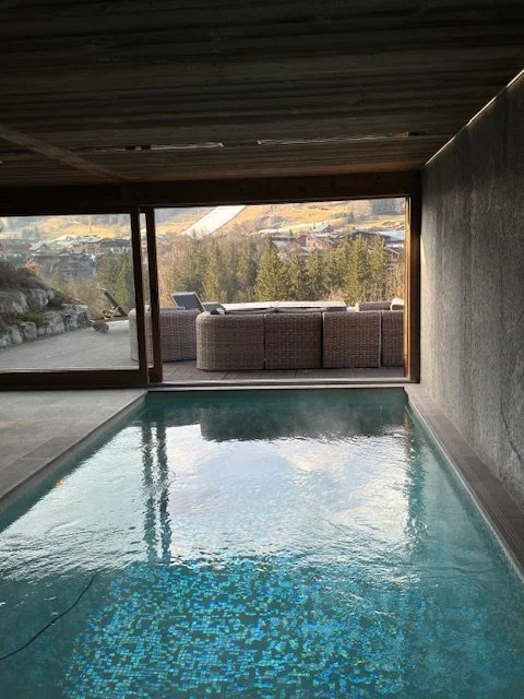 Indoor swimming pool with large windows overlooking an outdoor balcony and mountain landscape.