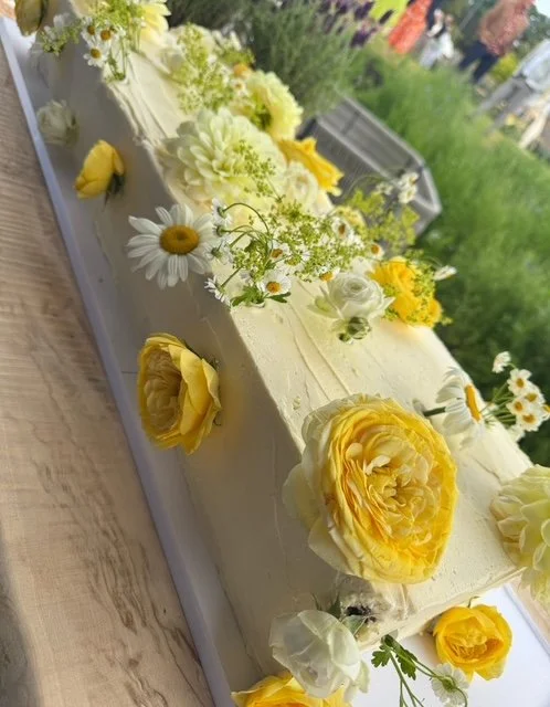 A long rectangular cake decorated with yellow and white flowers, including roses and daisies, in an outdoor setting with greenery in the background.