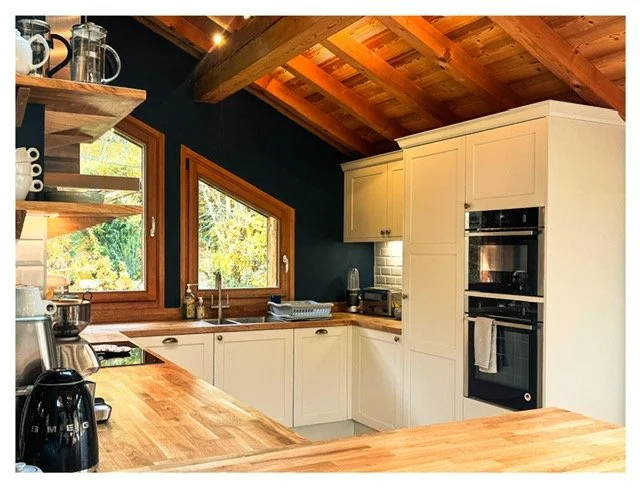 Kitchen with white cabinets, wooden countertops, and a dark accent wall, featuring large windows with wooden frames and a wooden ceiling with exposed beams.