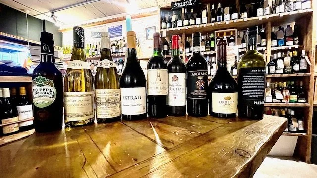 A variety of wine bottles and a green bottle on a wooden table inside a wine shop.