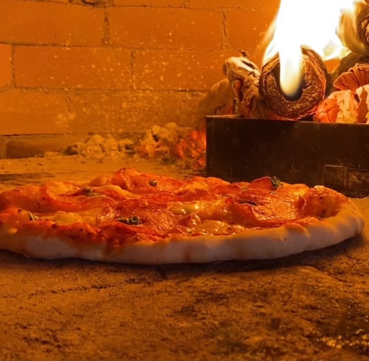 A pizza baking in a wood-fired oven with flames and burning wood logs in the background.