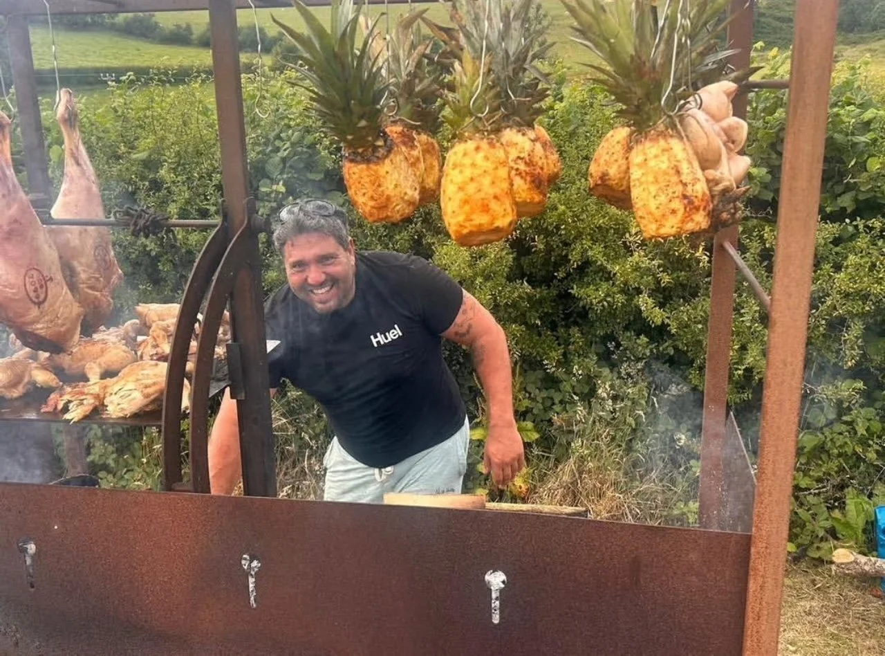 Man grilling meat and hanging pineapples, with a big smile on his face, outdoors in a grassy area with bushes in the background.