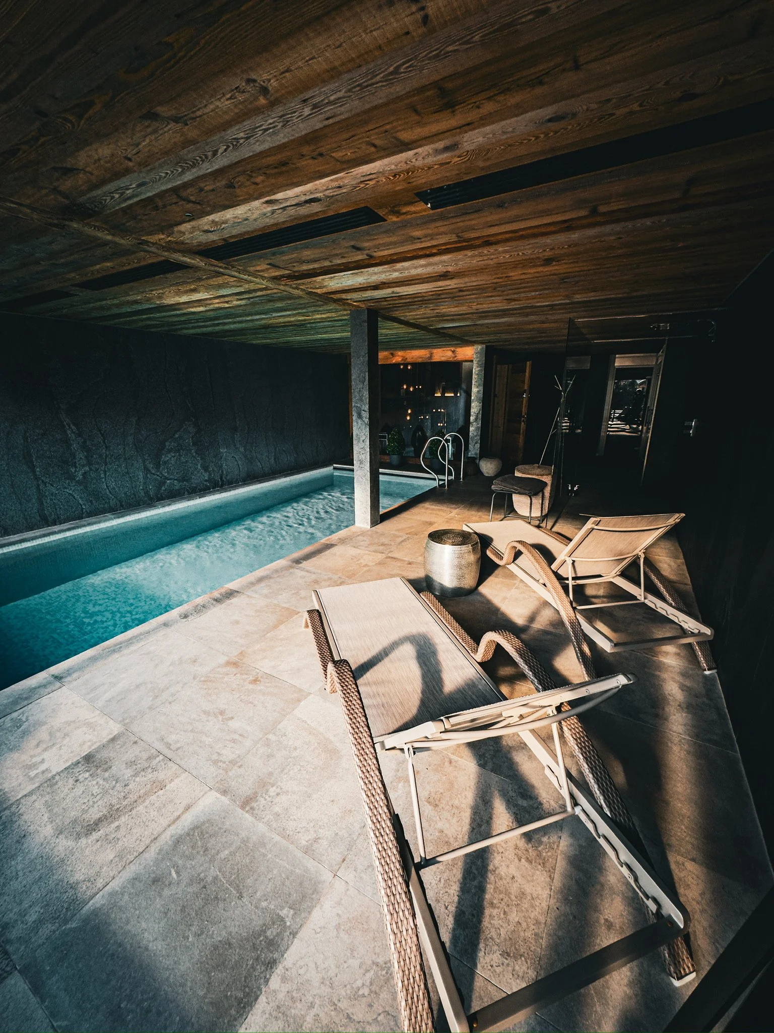 Indoor pool area with two lounge chairs, small table, and a textured wall, under a wooden ceiling.