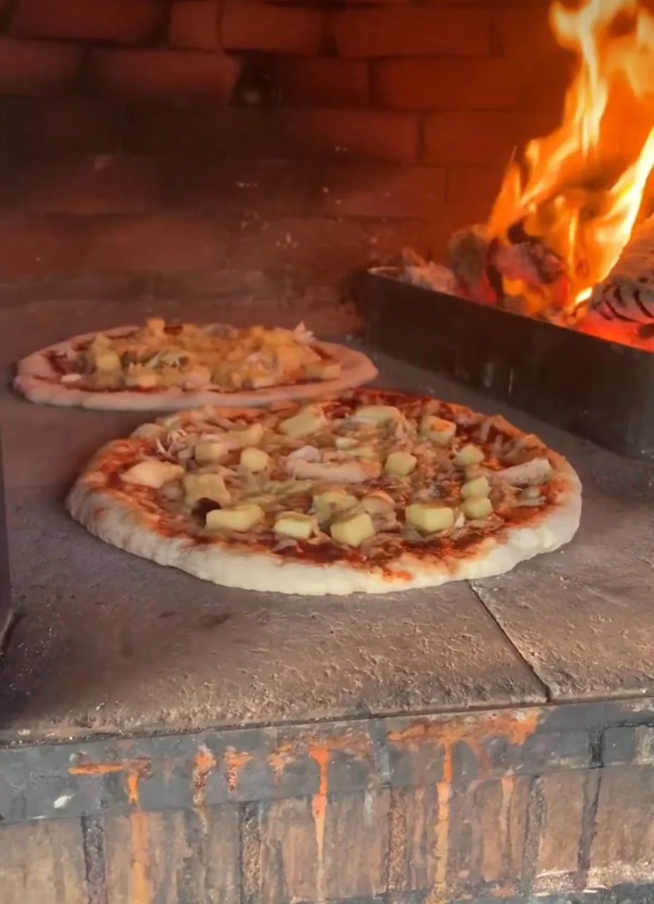 Two pizzas baking in a brick oven, flames visible in the background.