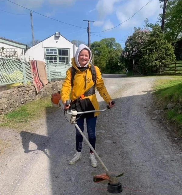 A woman outdoors on a gravel path, holding a string trimmer, wearing a yellow jacket, white hoodie, and backpack, with houses, trees, and a blue sky in the background.