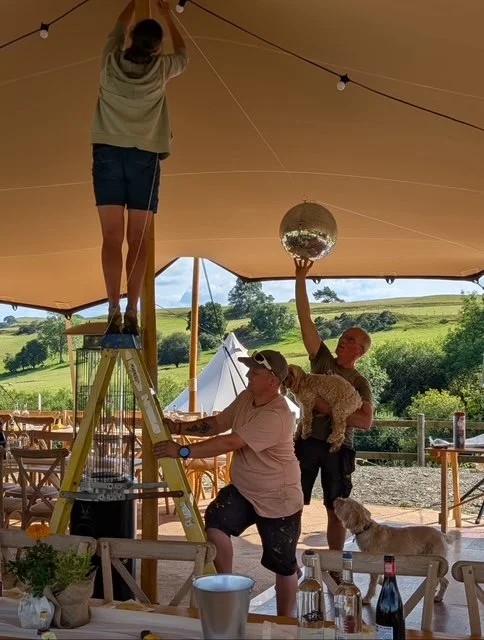 Three people are setting up decorations under a large outdoor tent by hanging a disco ball from the ceiling. One person is on a ladder securing the disco ball, while the other two are on the ground; one is holding the disco ball and the other is hold