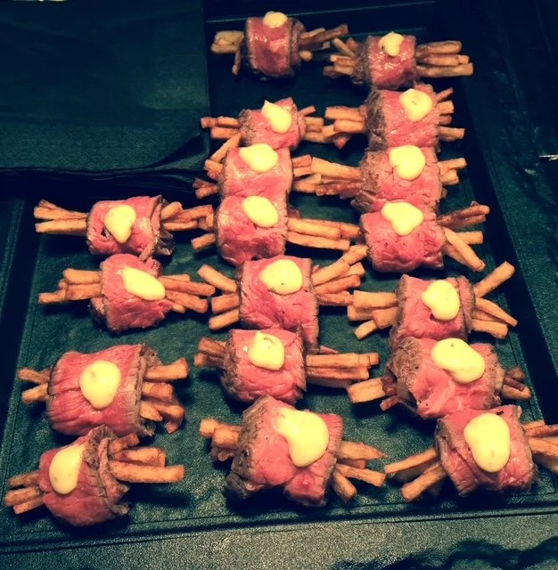 Bite-sized beef tenderloin topped with yellow sauce, wrapped around French fries on a black tray.