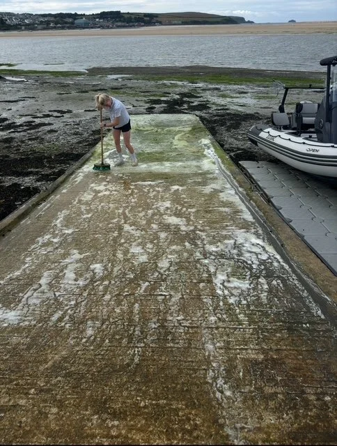 A girl cleaning a concrete dock with a broom near a boat by the water on a cloudy day.