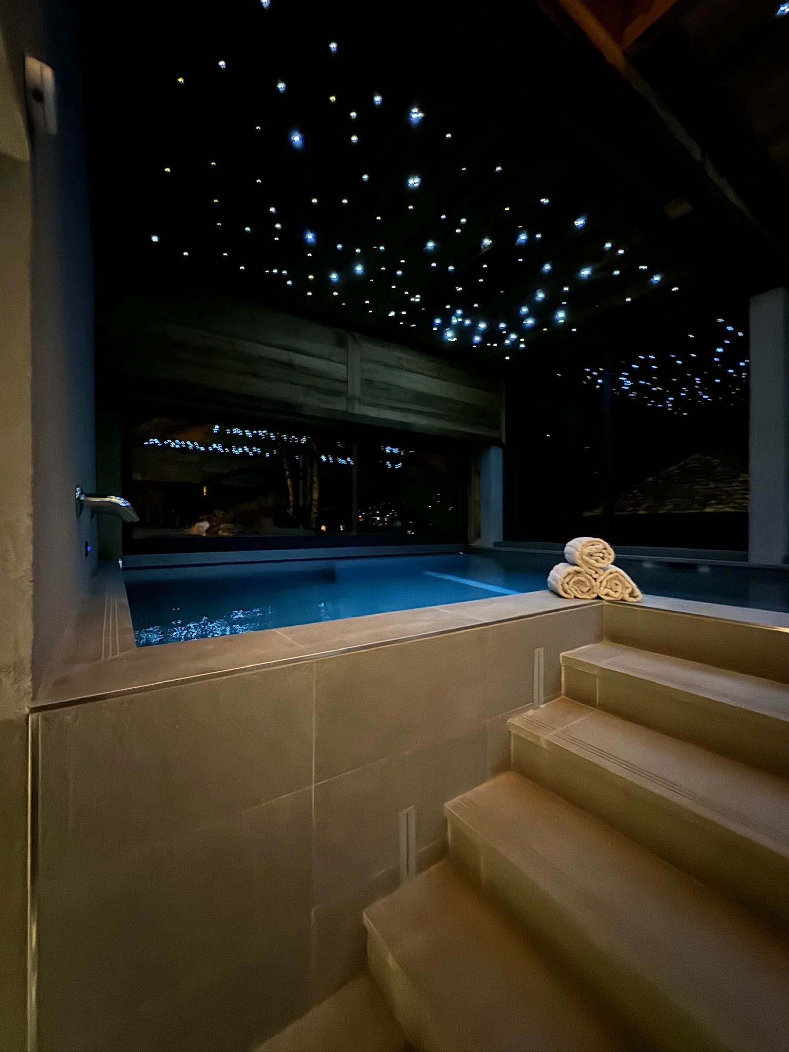 Indoor hot tub or pool with illuminated starry ceiling, folded towels on the side, and stairs leading up to the water, at night.