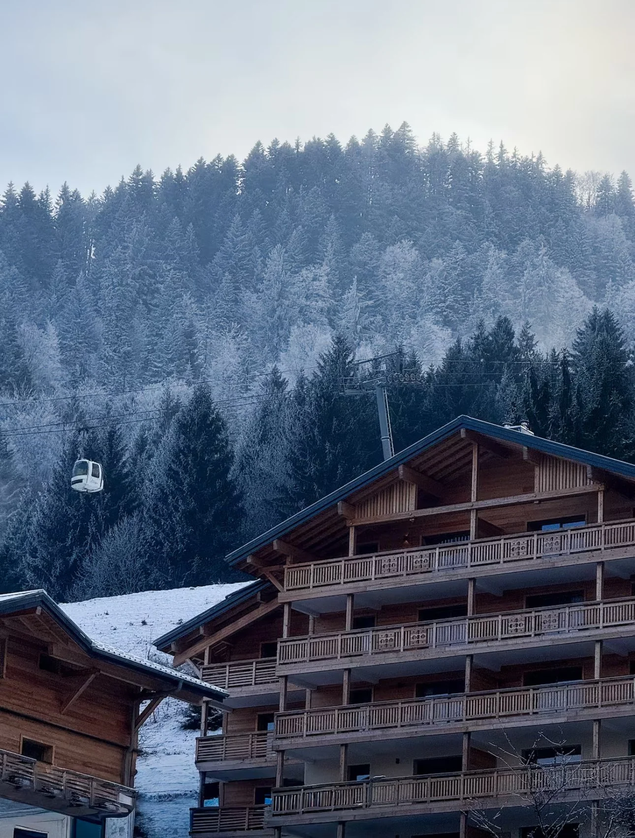 A winter scene with a wooden chalet-style building in the foreground, snow-covered ground, and a forested mountain with a cable car ascending in the background.
