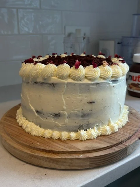 A round layered cake with white frosting on a wooden cake stand, decorated with white piped icing borders and topped with red and dark brown crumbs or flowers.