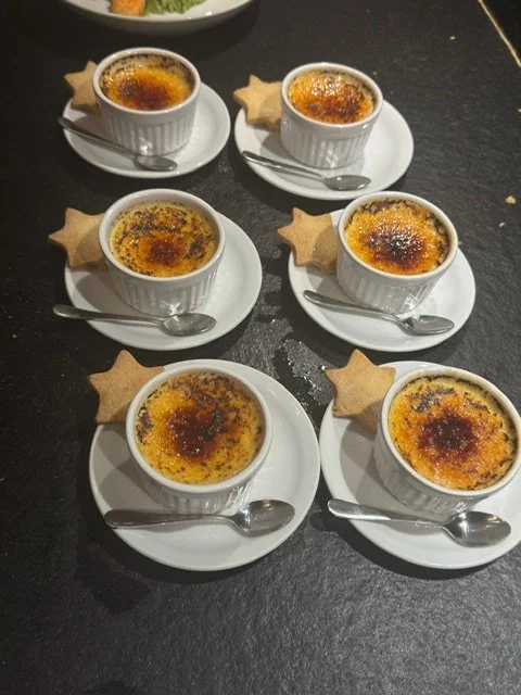 Six ramekins of crème brûlée on small plates with spoons, each topped with caramelized sugar, and decorated with star-shaped cookies.
