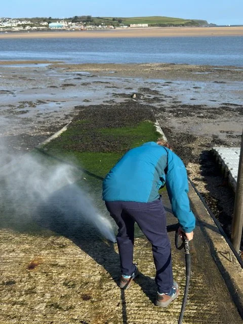A person in a blue jacket and dark pants pressure washing a concrete surface near a body of water on a sandy beach.