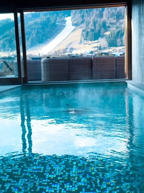 Indoor swimming pool with large window overlooking snow-covered mountains and trees.
