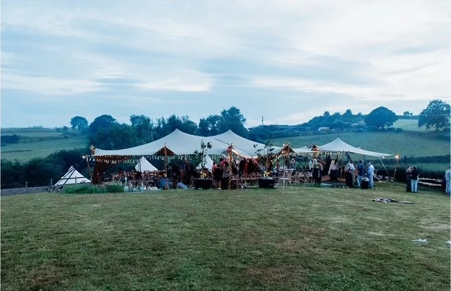 Outdoor event with large white tent set up in a grassy field during daytime, with people gathered around and a scenic rural landscape in the background.