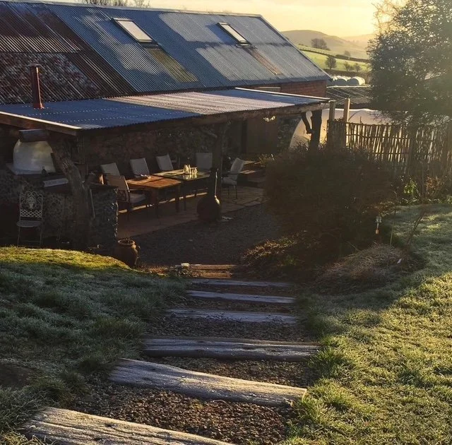 A rustic outdoor dining area with a long wooden table and chairs, under a metal roof attached to a house with solar panels, surrounded by a garden in the countryside at sunset.