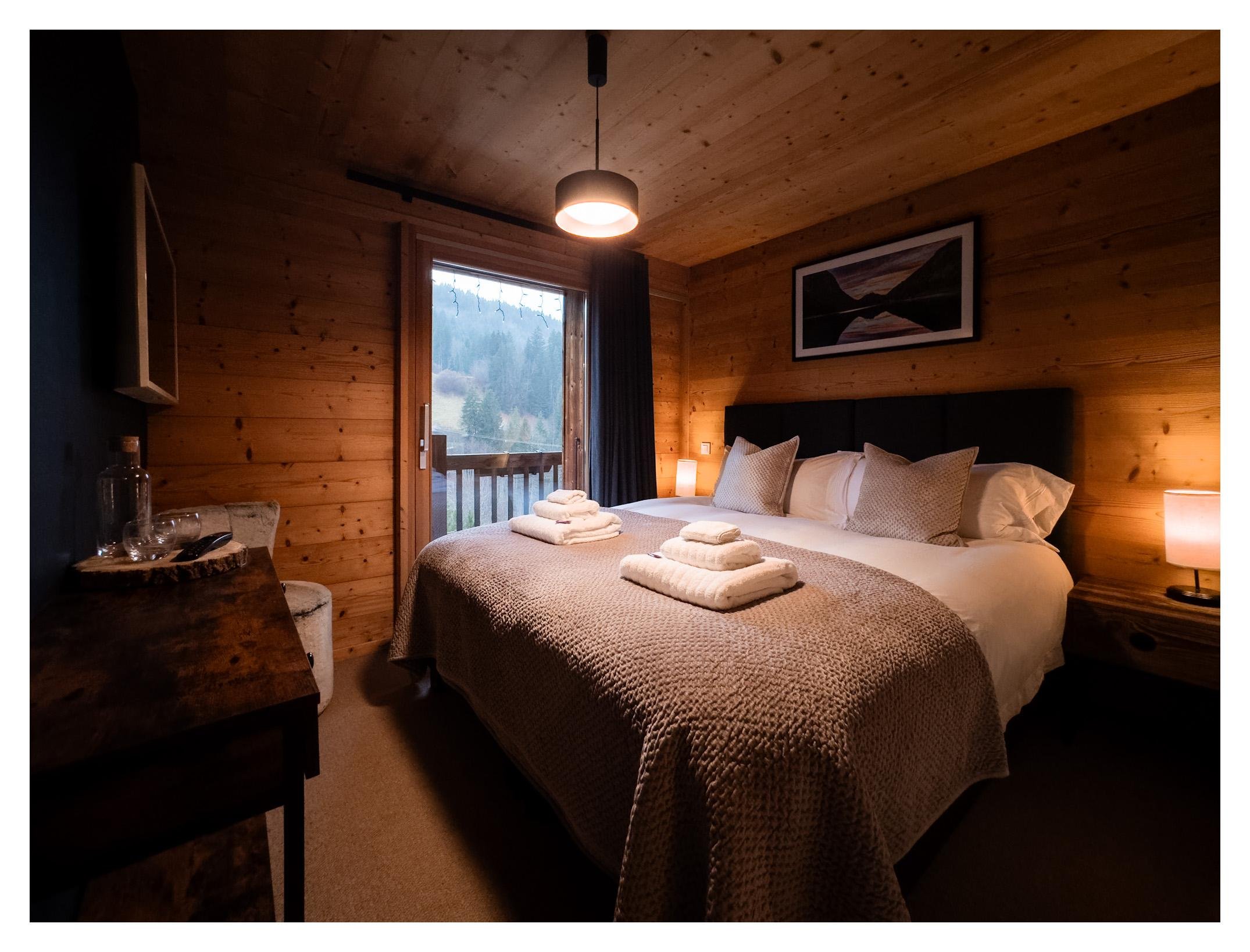 A cozy bedroom with wooden walls and ceiling, a double bed with white bedding and beige pillows, two folded towels on the bed, bedside tables with lamps, a framed landscape painting above the bed, a sliding glass door leading to a balcony with a moun