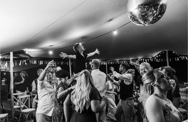 People dancing and celebrating at a party under a large tent, with a woman being lifted on someone's shoulders and a disco ball hanging from the ceiling.