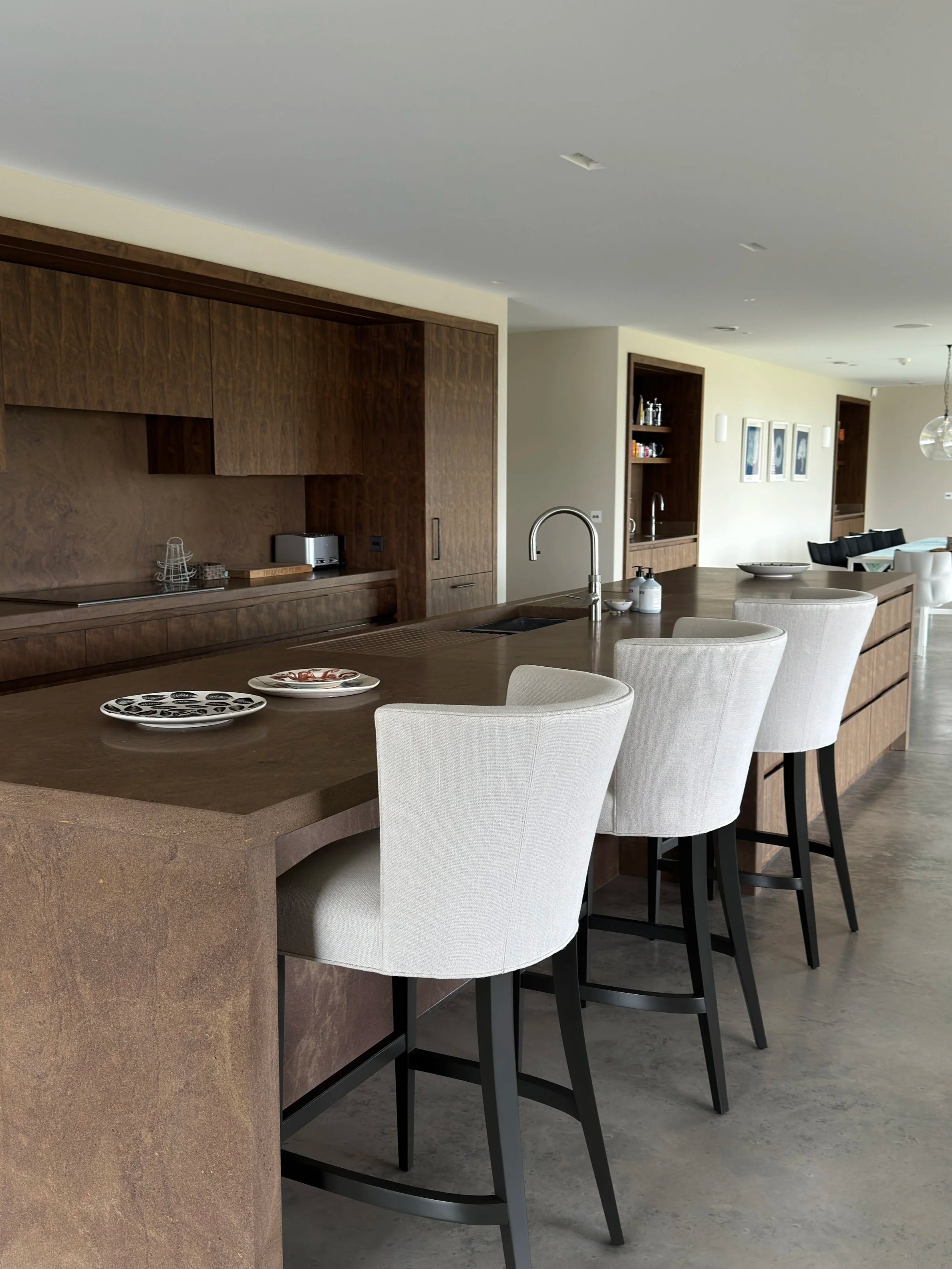 Modern kitchen with a large brown island, white upholstered bar stools, and built-in wooden cabinetry and shelving, with a view into a living area.