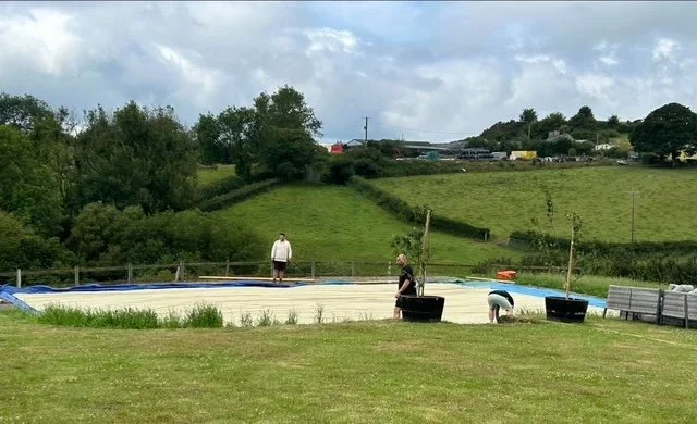 Three people are setting up a small outdoor swimming pool on a grassy field with rolling hills and trees in the background.