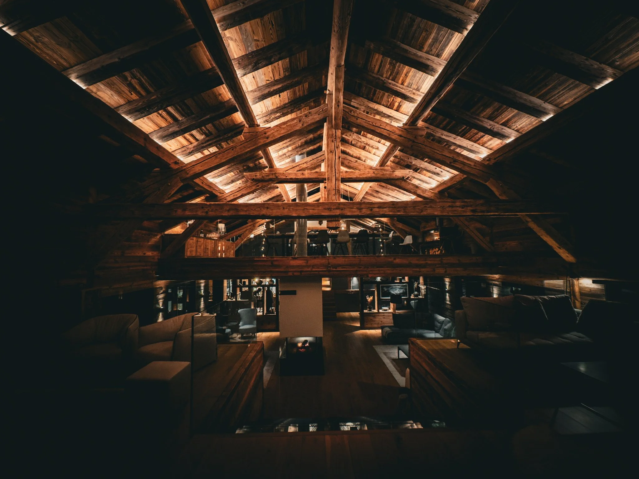 Interior view of a rustic wooden attic living space with exposed wooden beams and ceiling, furnished with sofas, chairs, and a fireplace.