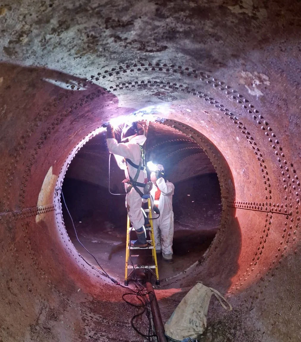 Workers in protective gear inspecting the interior of a large, rust-colored metal pipe, using flashlights and a ladder inside the pipe.