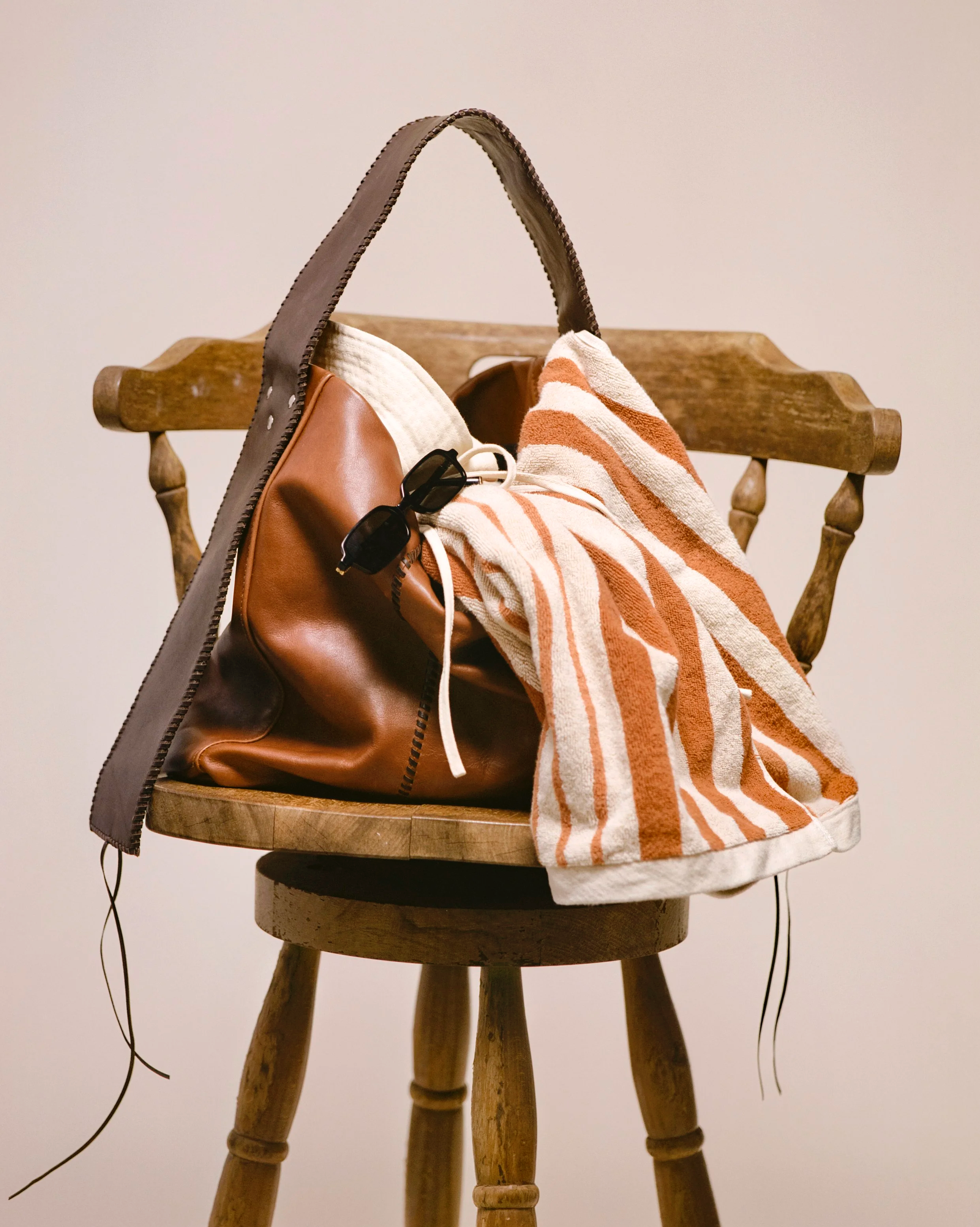 A wooden chair with a brown leather bag, a striped towel, a pair of sunglasses, and a pair of earphones on it.