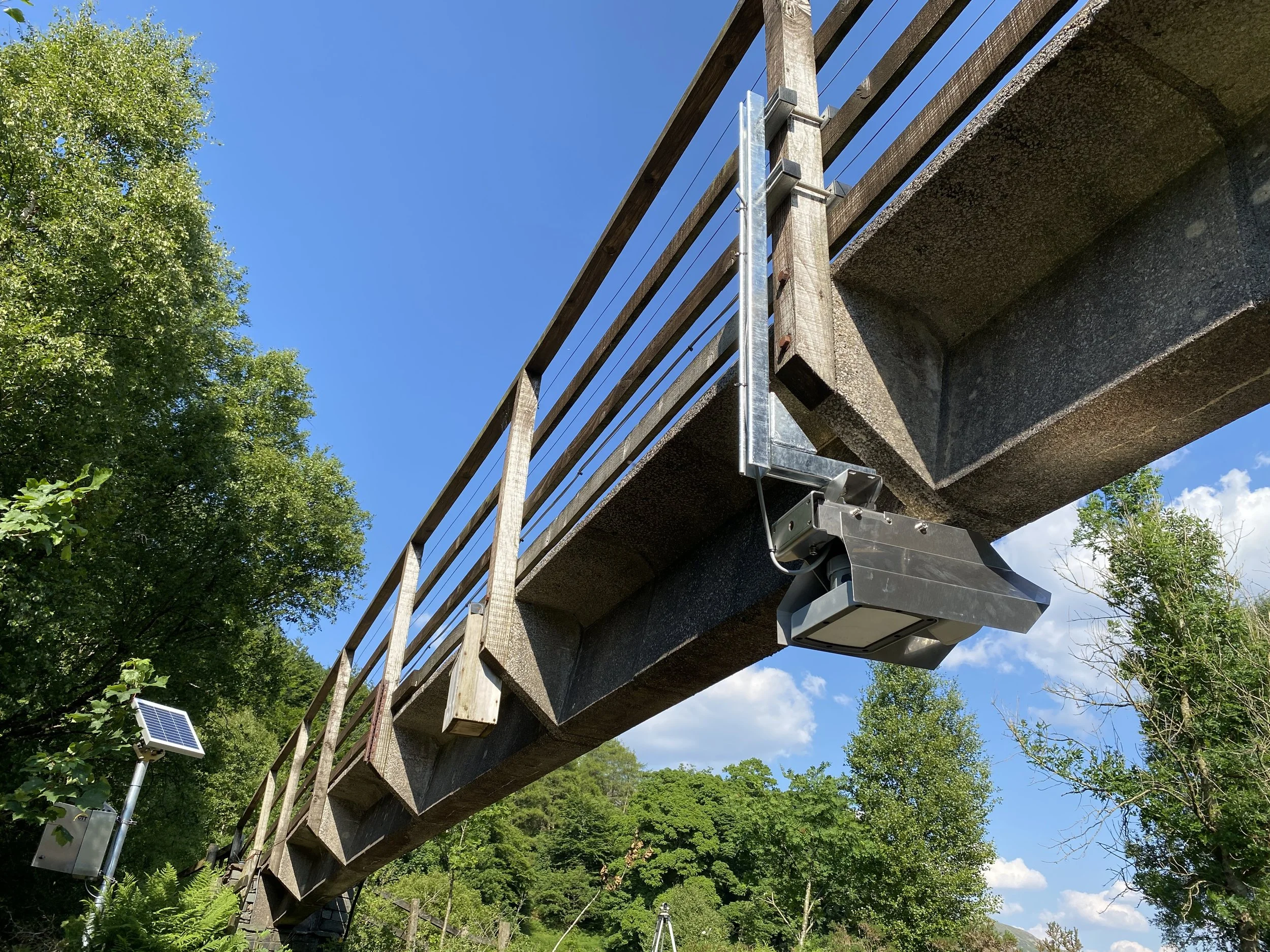 Underneath view of a bridge with a solar panel on a pole and electronic equipment attached to the underside, surrounded by green trees and a blue sky with clouds.