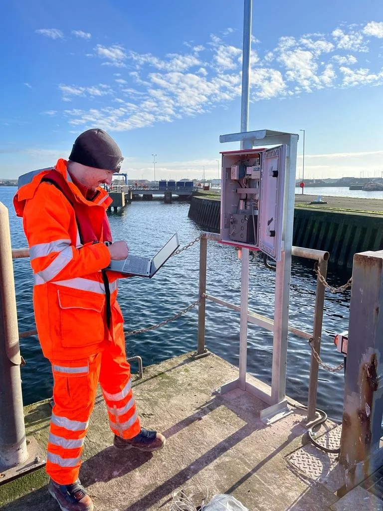 A person in an orange safety jacket and black beanie standing on a dock, working on a gray electrical box near the water, with a laptop in hand and a partly cloudy sky in the background.