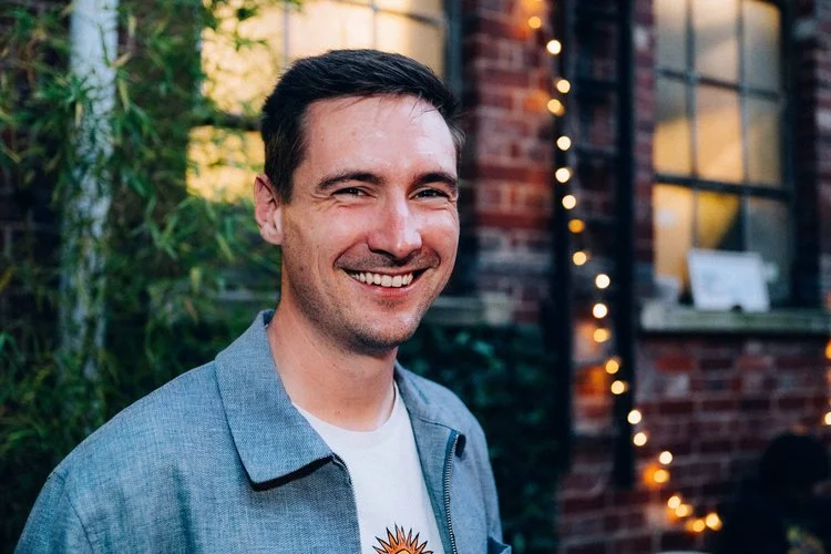 A smiling man with dark hair standing outdoors near a brick building decorated with string lights.