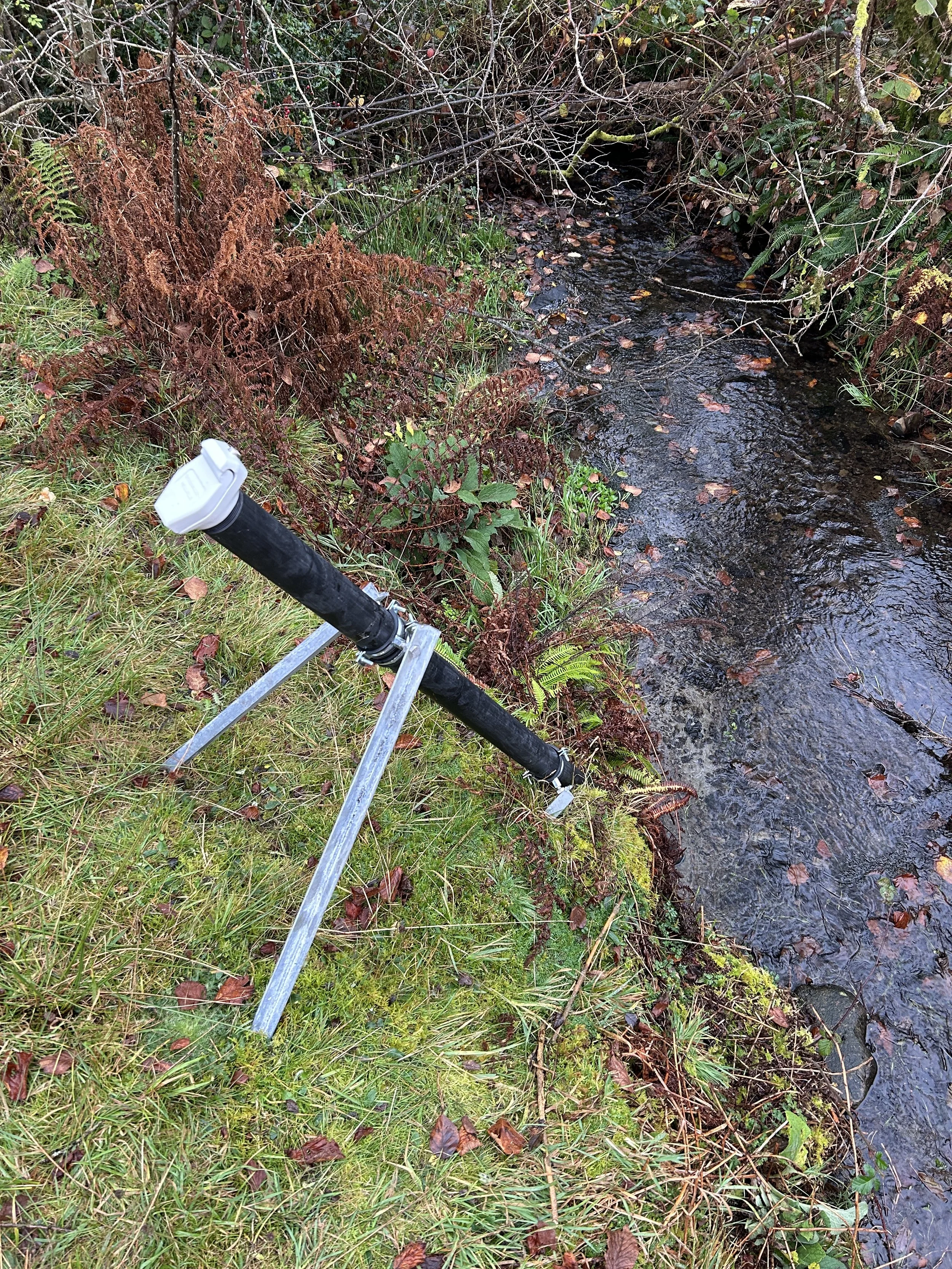 A portable pipe inspection camera on a tripod stand is set up on the grassy bank of a small stream surrounded by trees and ferns.