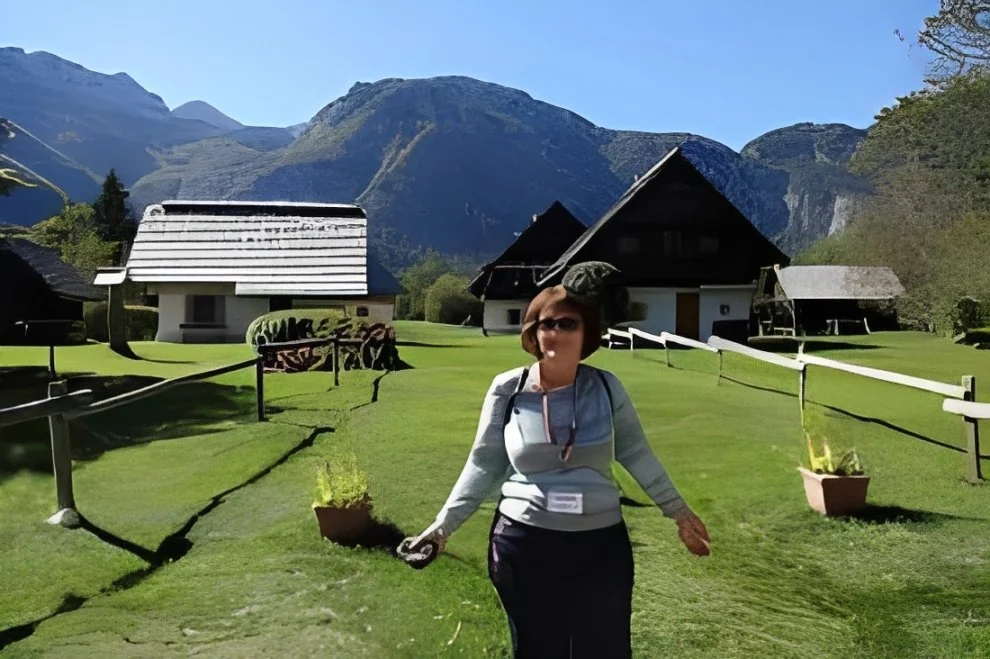 A woman stands on a grassy path in front of a scenic mountain landscape with traditional houses, under a clear blue sky.