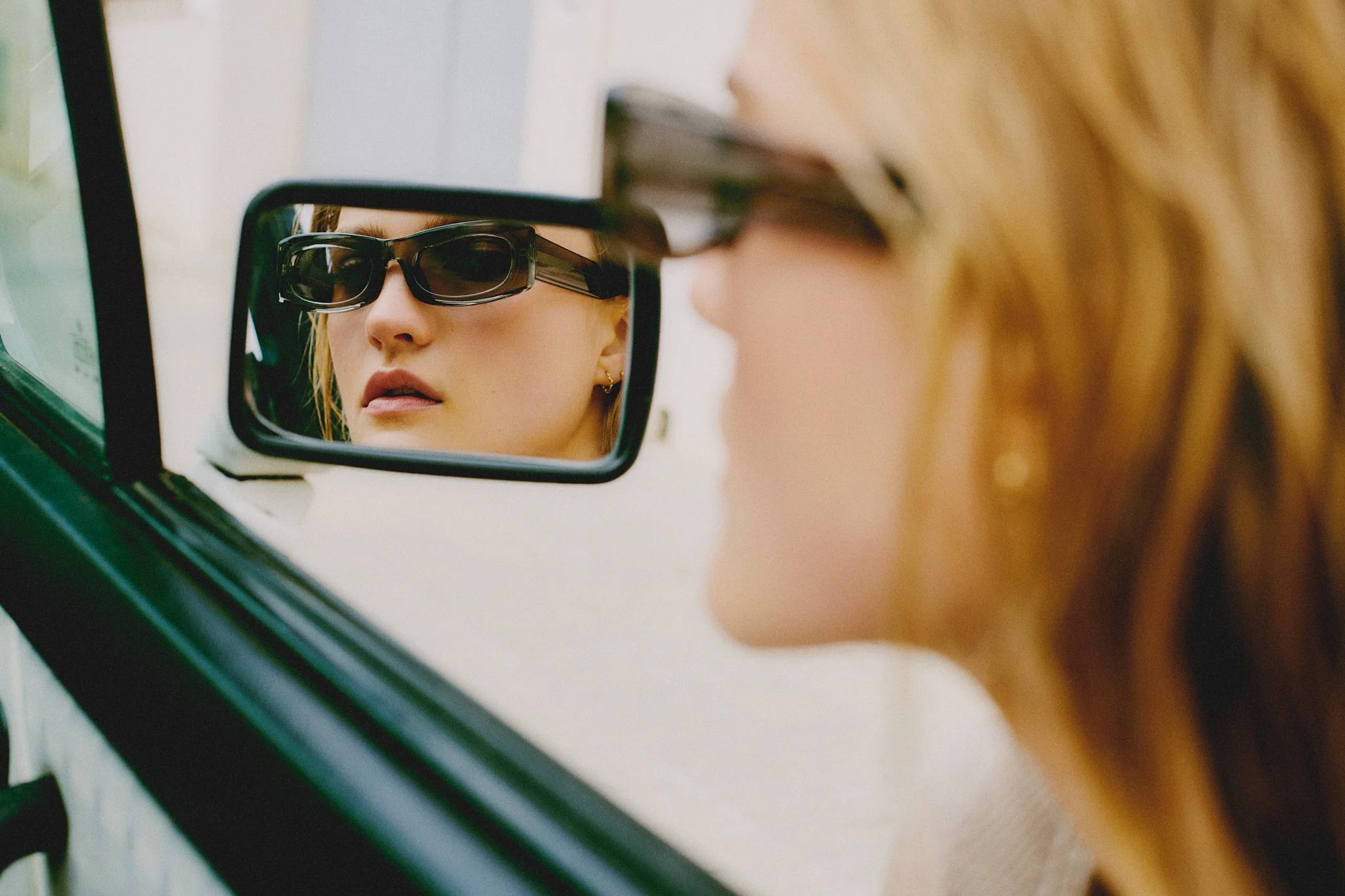 A woman with blonde hair wearing sunglasses looking into a rearview mirror, which reflects her face.