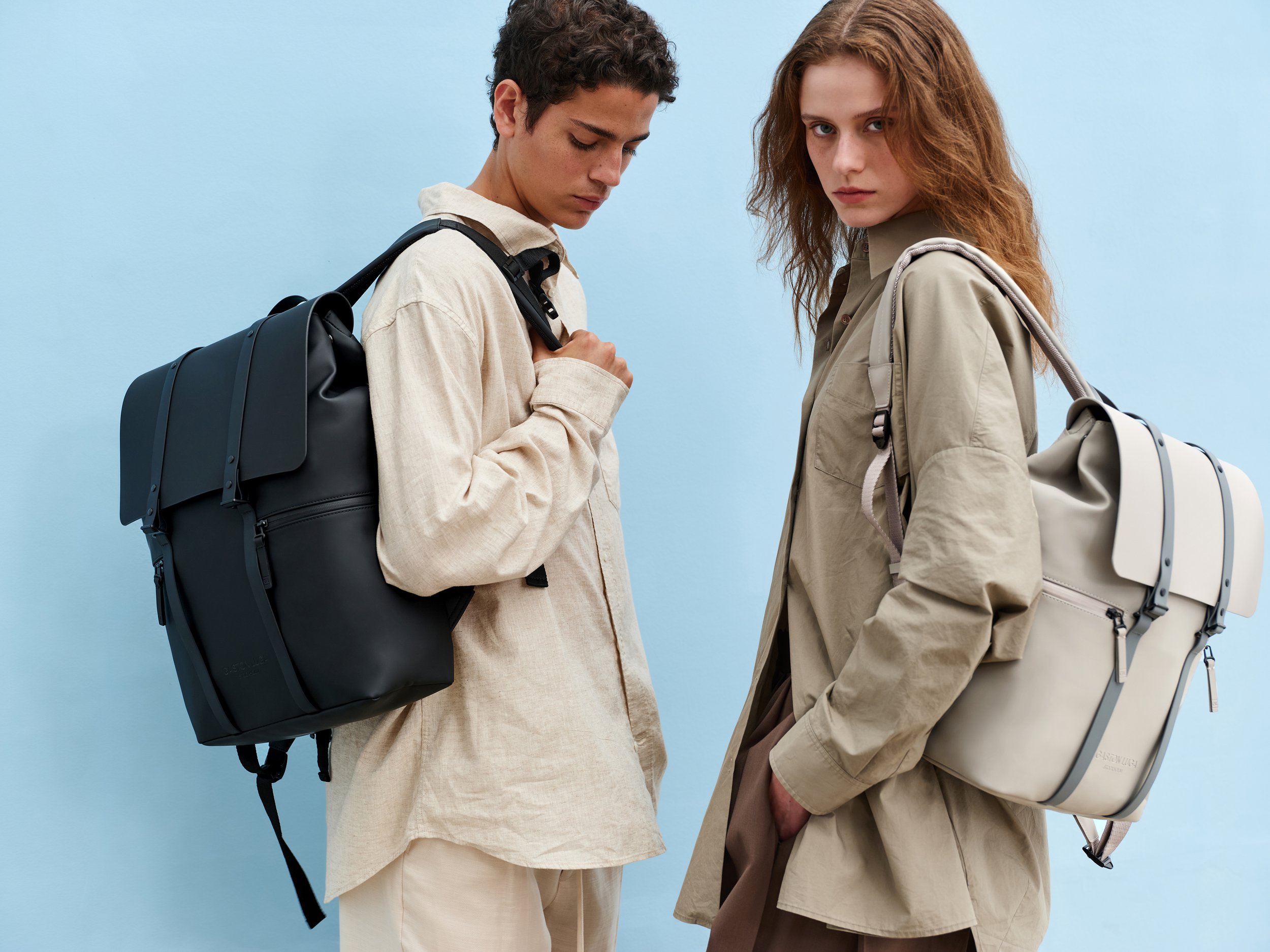 Two young adults, a man and a woman, standing against a light blue background, each carrying a large backpack and wearing beige and neutral-toned clothing.