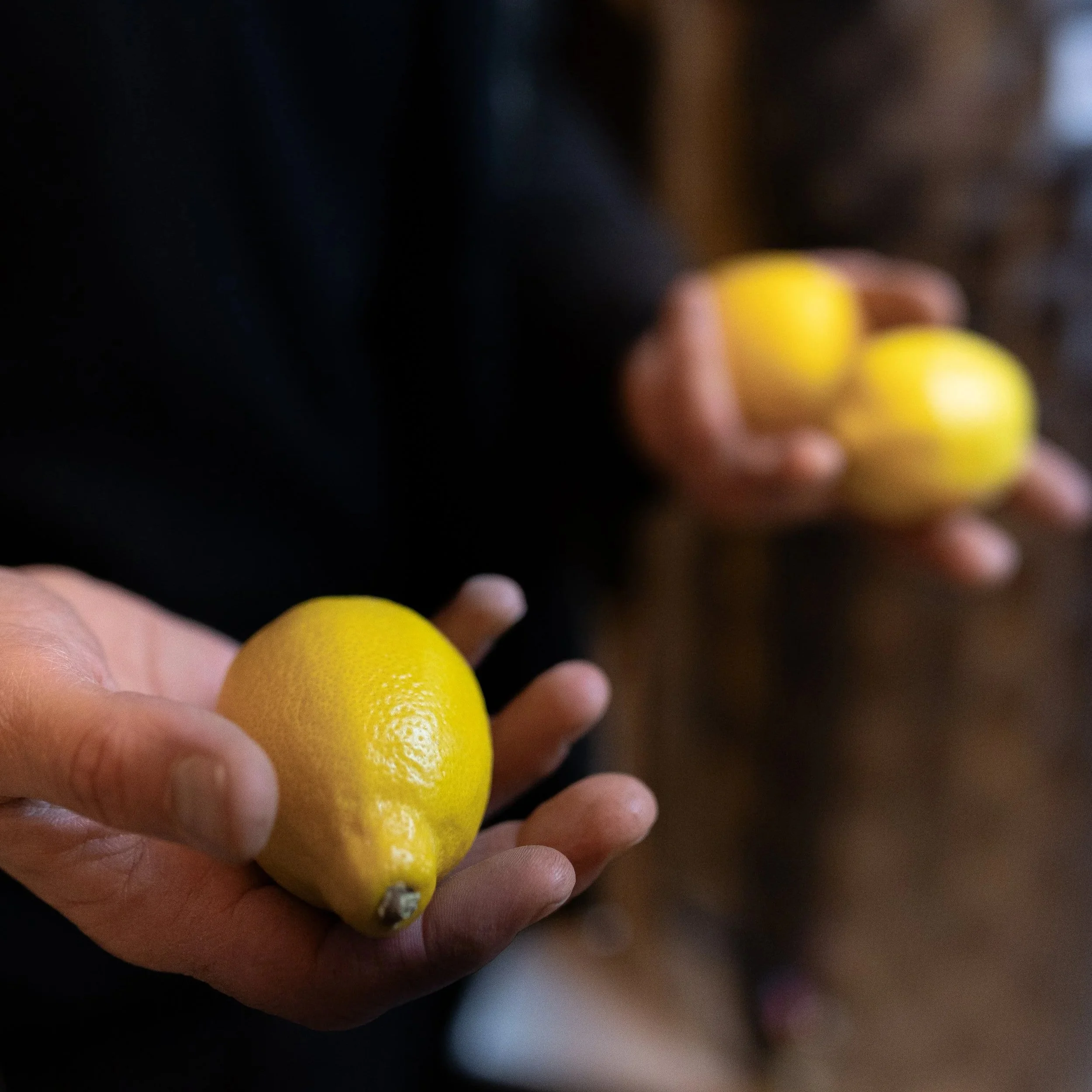Close-up of a person's hand holding a yellow lemon with a slightly curved shape and textured surface, with another person in the background holding three more yellow lemons.