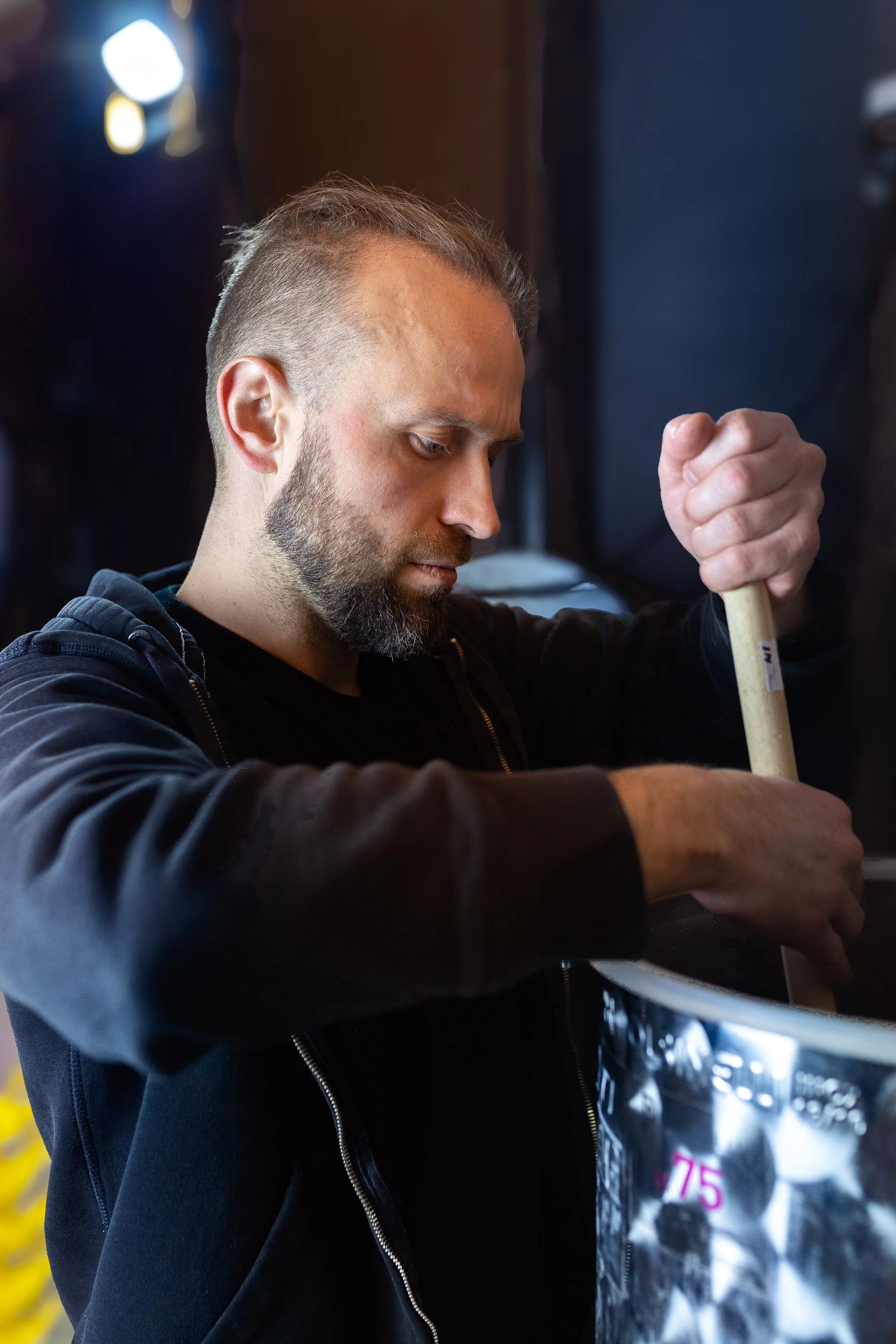 A man with short hair and a beard using a wooden tool to work on a metallic object in a workshop setting.