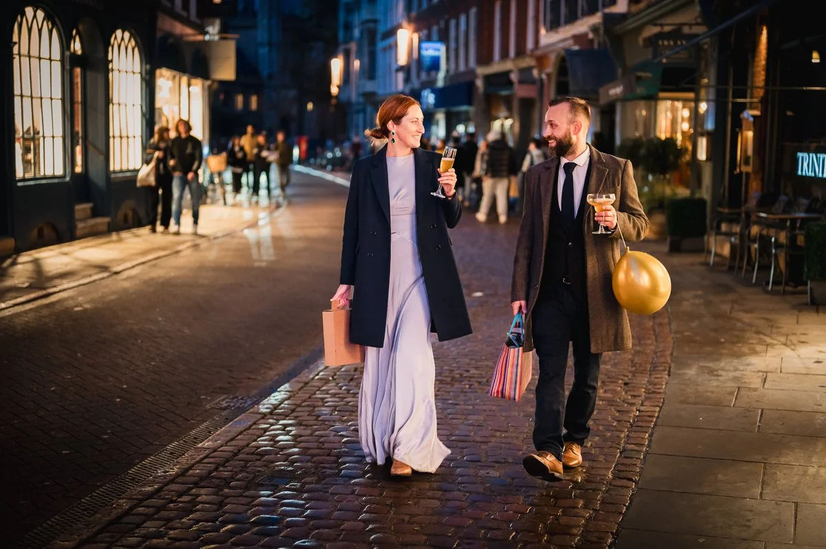 A man and woman walking down a cobblestone street at night, both holding drinks, smiling and talking, with shopping bags and holiday-themed balloons.
