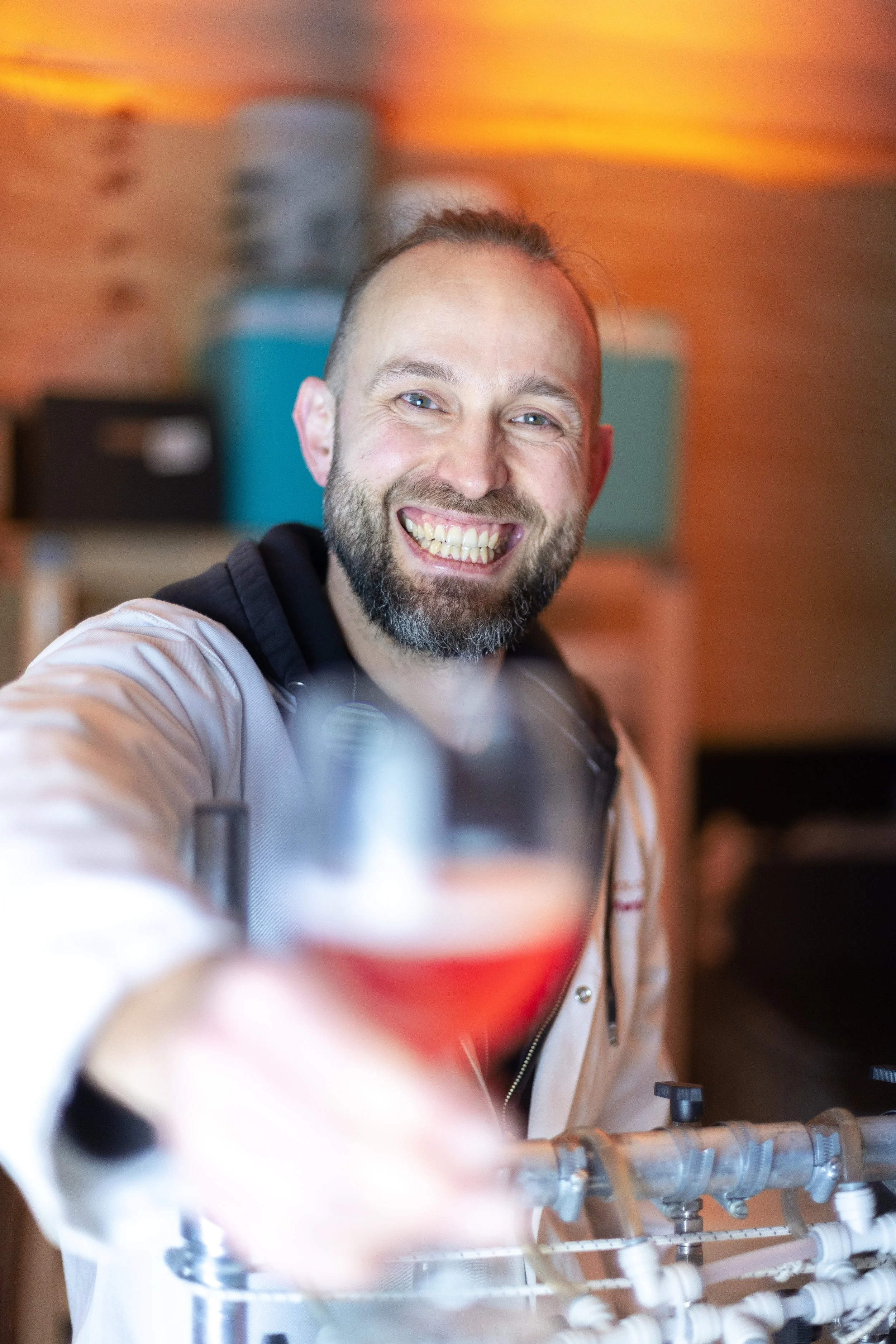 A smiling man in a light-colored jacket and hoodie holding a glass of red kombucha, with a blurred background.