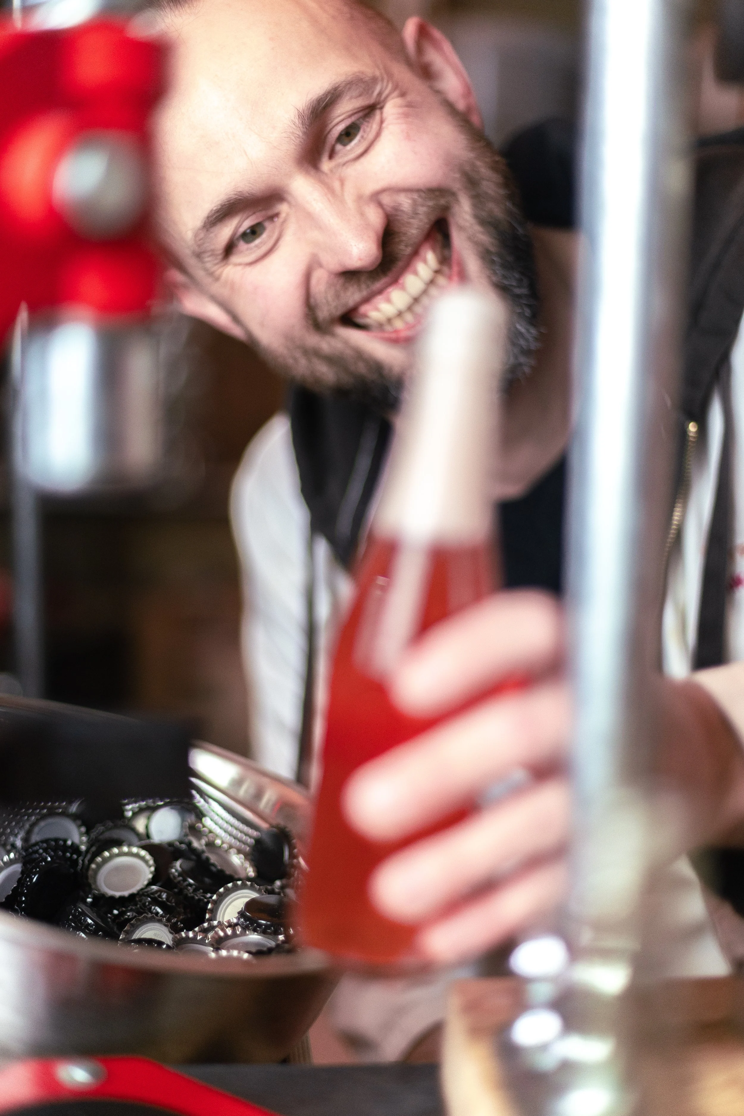 A man smiling and holding a red beverage bottle near a countertop with beverage caps or bottle caps.