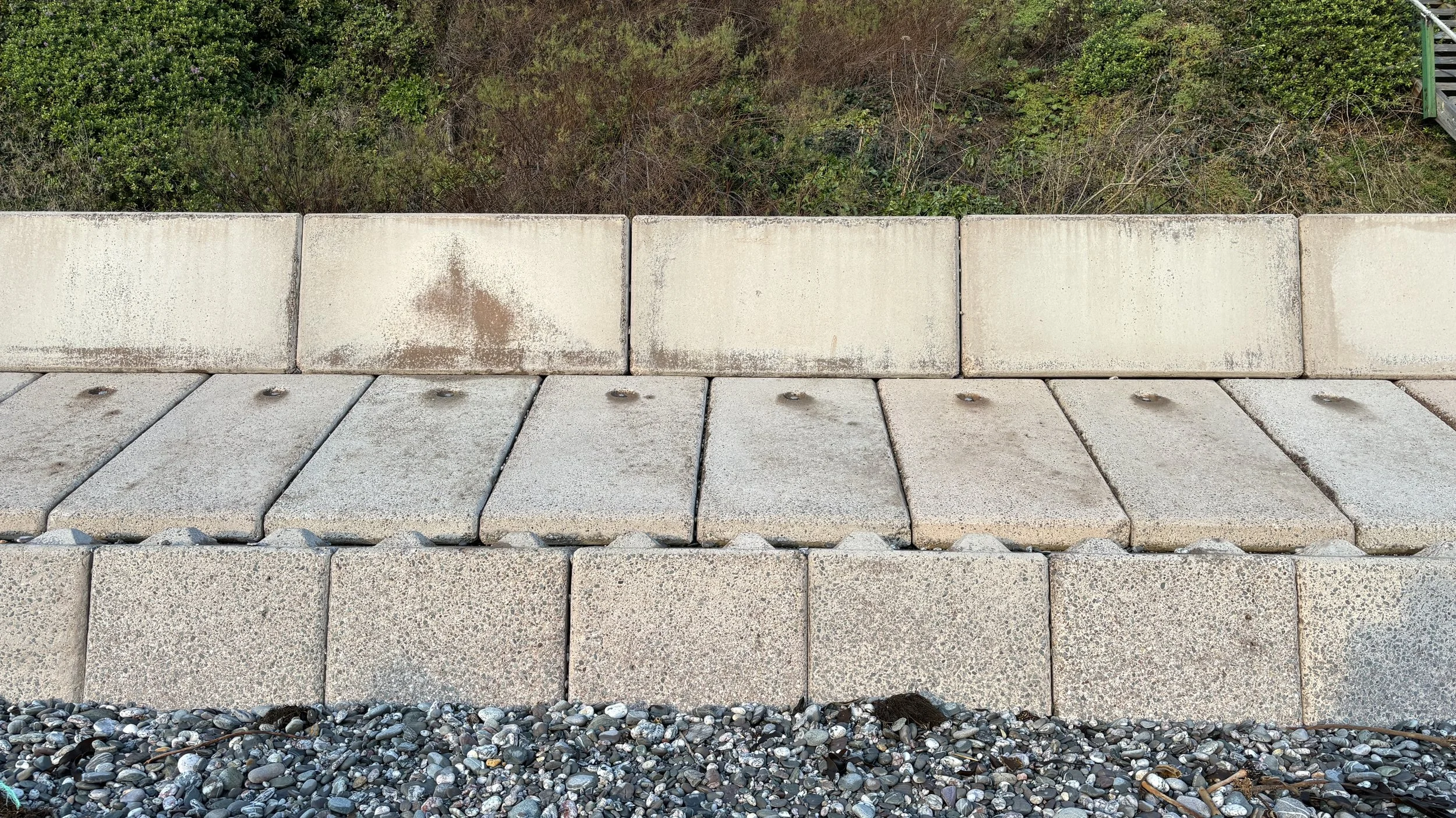 A straight-on view of a stone retaining wall with a garden in the background, consisting of large concrete blocks, with a layer of small rounded pebbles at the base.