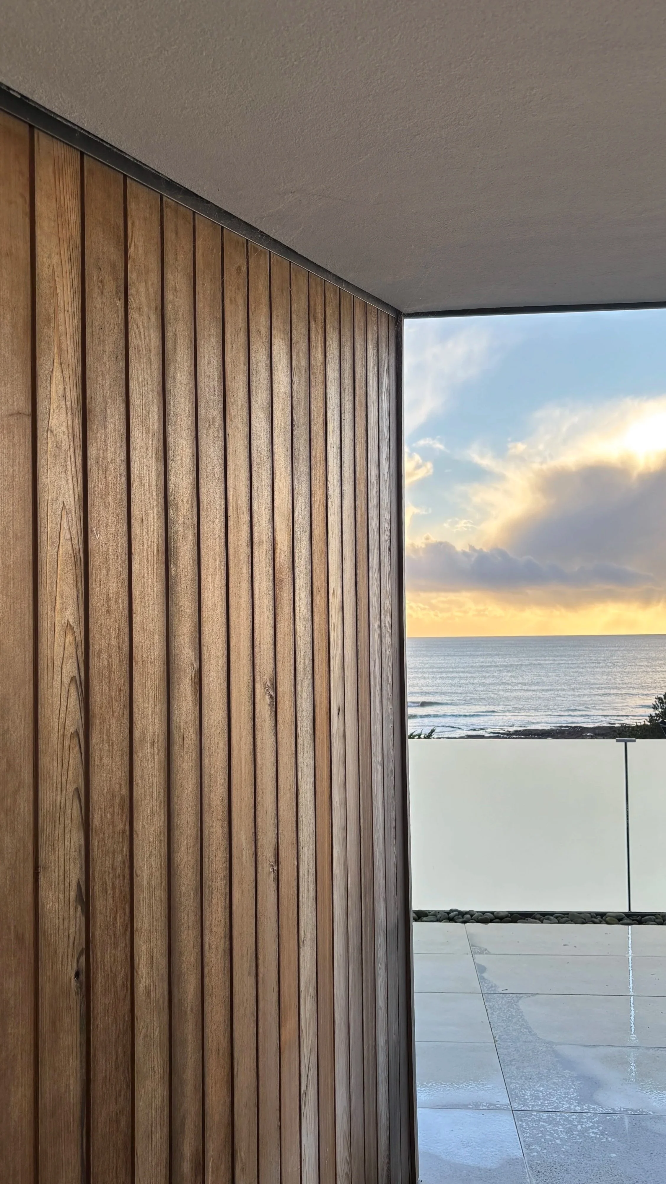 View from a balcony or patio overlooking the ocean at sunset, with a wooden wall on the left and a glass or clear railing.