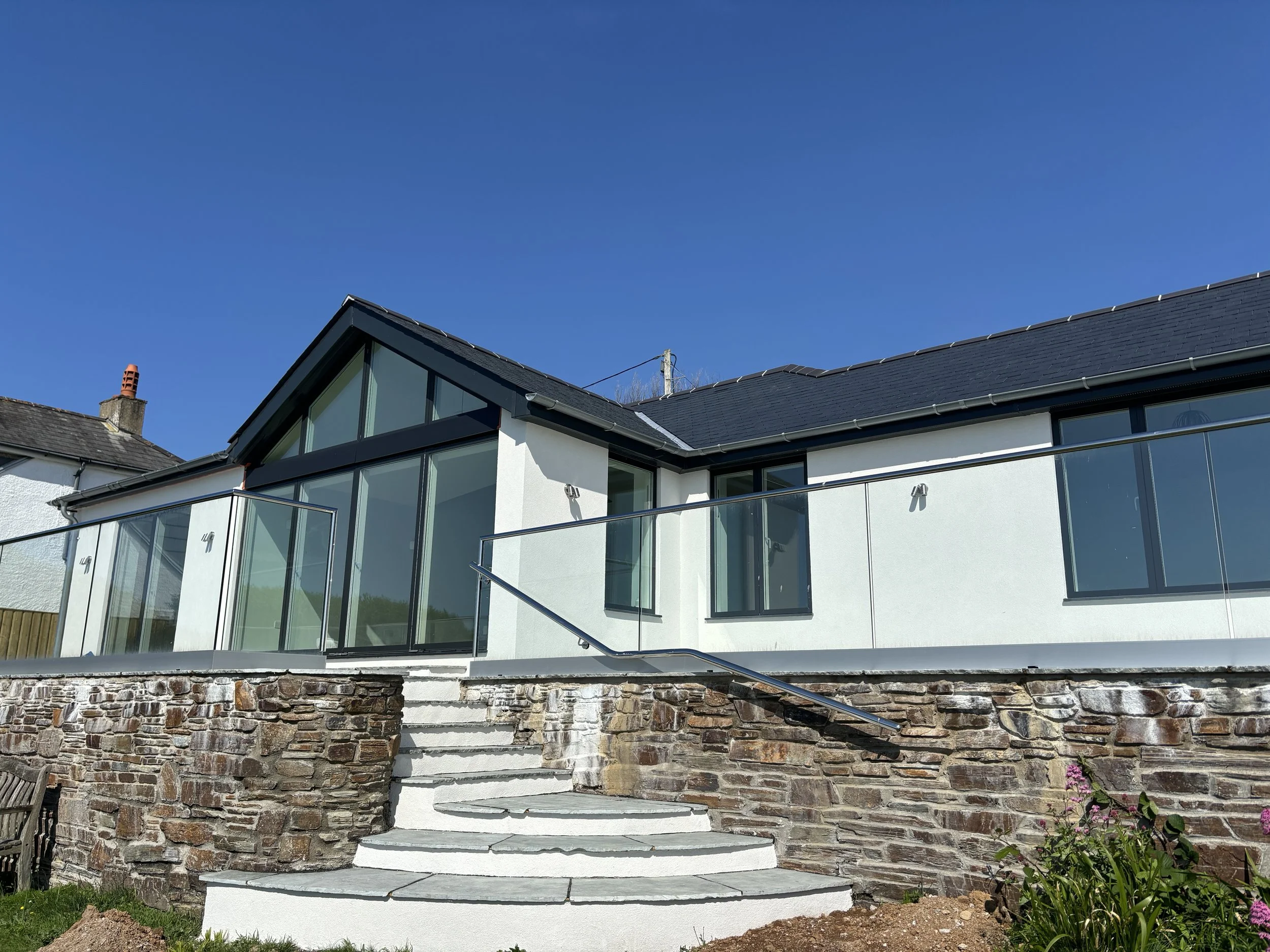 Modern house with white exterior, black roof, large glass windows and a glass balcony, stone stairs, and a landscaped garden with pink flowers.